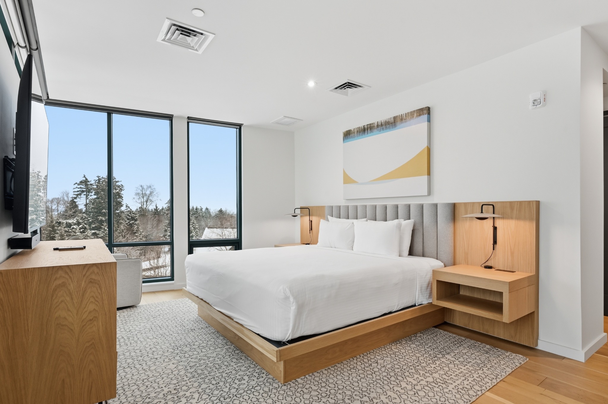 Bedroom with a low wood platform bed, built-in nightstands and floor-to-ceiling corner windows looking out to evergreens.